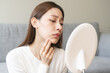 © KMPZZZ - Expression worry asian young woman hand touching pustule around the chin and mouth, allergic when wear mask, makeup, show squeezing pimple spot from face. Beauty care, skin problem by acne treatment.
