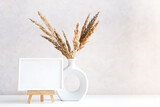Fototapeta Boho - Empty picture frame mock up in minimalist interior with dried plant in vase on white table.