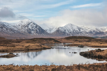 Wall Mural - Majestic Winter panorama landscape image of mountain range and peaks viewed from Loch Ba in Scottish Highlands with dramatic clouds overhead