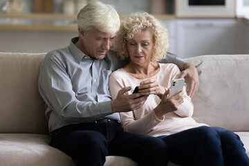 Serious elderly 70s couple sit on sofa hold two new bought modern smartphones, try to understand mobile application discussing software. Older generation use wireless gadgets, technology, lifestyle