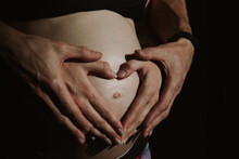 Pregnant Woman With Support Of Her Man. Hands Of A Man And Woman Showing Heart Around The Belly Button. Couple In Love Expecting Baby. 