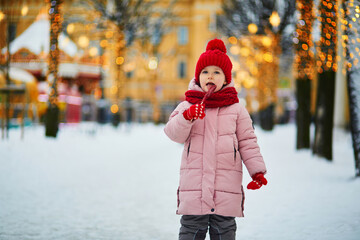 Wall Mural - Happy preschooler girl in red snood and hat having fun on a Christmas market and eating sugar candy