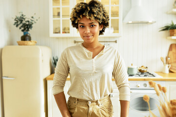 Wall Mural - Cute happy youthful girl of 20s with dark skin and afro hair looking at camera with shy face posing on kitchen background against fridge in stylish casual clothes, going to eat something sweet