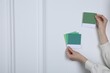 Woman with color sample cards choosing paint shade near white wall, closeup. Interior design