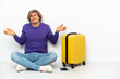 © luismolinero - Young blonde man with suitcase sitting on the floor having doubts while raising hands