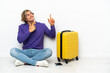 © luismolinero - Young blonde man with suitcase sitting on the floor pointing with the index finger a great idea