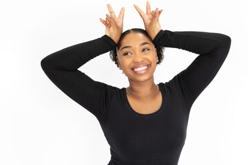 Wall Mural - Portrait of cheerful young woman making bunny ears gesture. African American lady wearing black longsleeve looking away and smiling against white background. Humor concept