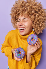 Wall Mural - Overjoyed curly haired woman laughs joyfully holds two delicious doughnuts happy to eat favorite dessert keeps eyes closed wears yellow jumper isolated over purple background. Sweet food concept