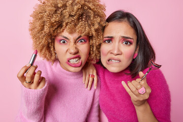 Photo of two irritated female models look angrily cannot apply professional makeup use professional cosmetics hold lipstick eyelashes curler angry to have spoiled makeup isolated on pink background