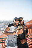 Fototapeta Miasto - Portrait of happy African gay couple taking selfie together. Two bearded men in casual clothes standing close on balcony hugging laughing looking at phone camera. LGBT peoples love, happiness concept