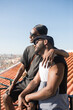 © KAMPUS - Side view of African gay couple admiring city view. Two lovers in sunglasses dating on roof top. One man sitting on fence hugging another man both looking far. Same sex couples happy relations concept