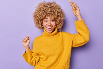 Wall Mural - Waist up shot of overjoyed curly haired dances carefree holds mobile phone moves rhythm of music dressed in casual yellow jumper isolated over purple background. People technology and fun concept