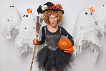 Wall Mural - Annoyed curly haired woman frowns face poses with broom and pumpkin wears dress and hat with autumn leaves isolated over grey background with mysterious ghosts around. Happy Halloween concept