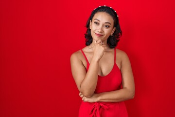 Wall Mural - Young hispanic woman standing over red background looking confident at the camera smiling with crossed arms and hand raised on chin. thinking positive.