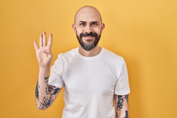 Sticker - Young hispanic man with tattoos standing over yellow background showing and pointing up with fingers number four while smiling confident and happy.