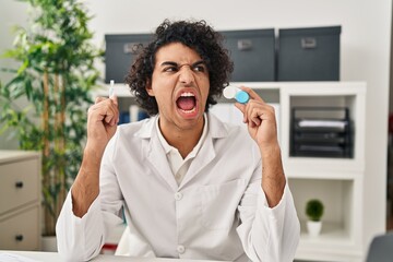 Poster - Hispanic man with curly hair holding contact lenses angry and mad screaming frustrated and furious, shouting with anger. rage and aggressive concept.