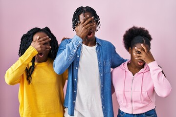 Canvas Print - Group of three young black people standing together over pink background peeking in shock covering face and eyes with hand, looking through fingers with embarrassed expression.