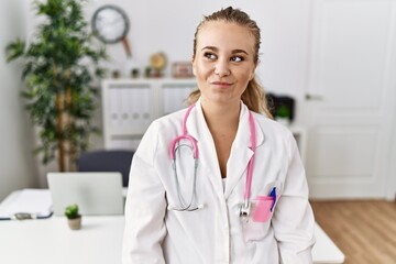 Wall Mural - Young caucasian woman wearing doctor uniform and stethoscope at the clinic smiling looking to the side and staring away thinking.