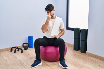 Wall Mural - Hispanic man with beard sitting on pilate balls at yoga room smelling something stinky and disgusting, intolerable smell, holding breath with fingers on nose. bad smell