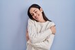 © Krakenimages.com - Young brunette woman standing over blue background hugging oneself happy and positive, smiling confident. self love and self care