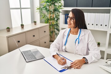 Poster - Young hispanic woman wearing doctor uniform writing on clipboard at clinic