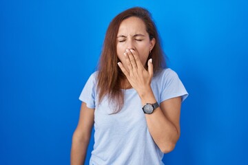 Poster - Brunette woman standing over blue background bored yawning tired covering mouth with hand. restless and sleepiness.