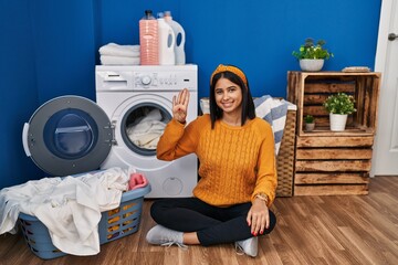 Poster - Young hispanic woman doing laundry showing and pointing up with fingers number four while smiling confident and happy.