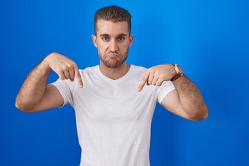 Poster - Young caucasian man standing over blue background pointing down looking sad and upset, indicating direction with fingers, unhappy and depressed.