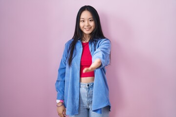 Young asian woman standing over pink background smiling friendly offering handshake as greeting and welcoming. successful business.