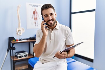 Poster - Young hispanic man wearing physiotherapist uniform talking on the smartphone at clinic