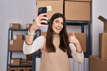 Wall Mural - Young brunette woman working at small business ecommerce taking selfie smiling happy and positive, thumb up doing excellent and approval sign