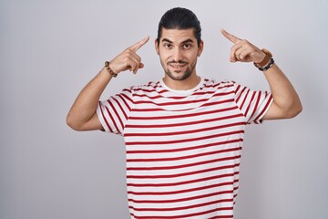 Wall Mural - Hispanic man with long hair standing over isolated background smiling pointing to head with both hands finger, great idea or thought, good memory