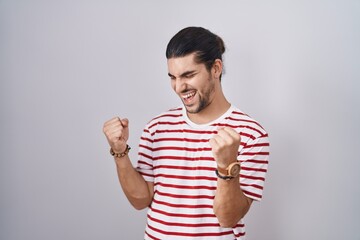 Wall Mural - Hispanic man with long hair standing over isolated background very happy and excited doing winner gesture with arms raised, smiling and screaming for success. celebration concept.