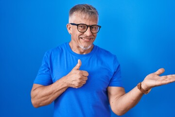 Canvas Print - Hispanic man with grey hair standing over blue background showing palm hand and doing ok gesture with thumbs up, smiling happy and cheerful