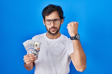 Canvas Print - Handsome latin man holding dollars banknotes annoyed and frustrated shouting with anger, yelling crazy with anger and hand raised