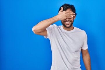 Poster - Handsome latin man standing over blue background smiling and laughing with hand on face covering eyes for surprise. blind concept.