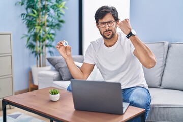 Poster - Handsome latin man holding virtual currency bitcoin using laptop annoyed and frustrated shouting with anger, yelling crazy with anger and hand raised