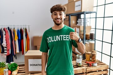 Wall Mural - Young arab man wearing volunteer t shirt at donations stand doing happy thumbs up gesture with hand. approving expression looking at the camera showing success.