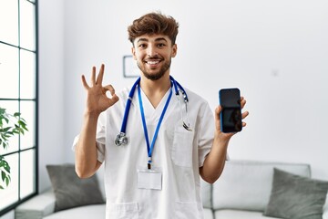 Wall Mural - Young arab man wearing doctor uniform and stethoscope holding smartphone doing ok sign with fingers, smiling friendly gesturing excellent symbol