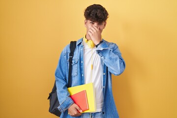 Hispanic teenager wearing student backpack and holding books smelling something stinky and disgusting, intolerable smell, holding breath with fingers on nose. bad smell
