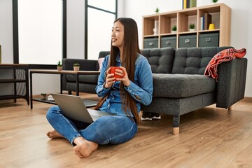 Wall Mural - Young chinese girl using laptop and drinking coffee sitting on the floor at home.