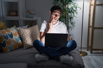 Poster - Young hispanic man using laptop at home at night touching mouth with hand with painful expression because of toothache or dental illness on teeth. dentist concept.