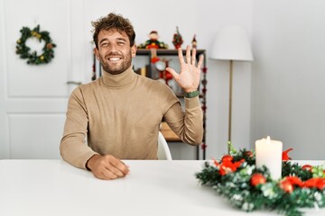 Sticker - Young handsome man with beard sitting on the table by christmas decoration showing and pointing up with fingers number five while smiling confident and happy.