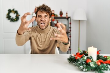 Wall Mural - Young handsome man with beard sitting on the table by christmas decoration shouting frustrated with rage, hands trying to strangle, yelling mad