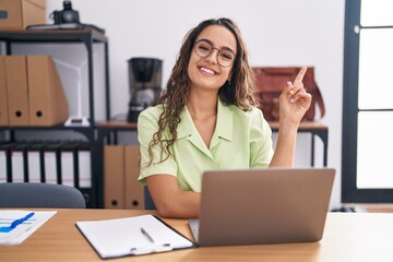 Wall Mural - Young hispanic woman working at the office wearing glasses with a big smile on face, pointing with hand and finger to the side looking at the camera.