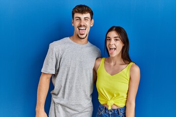 Poster - Young hispanic couple standing together over blue background sticking tongue out happy with funny expression. emotion concept.