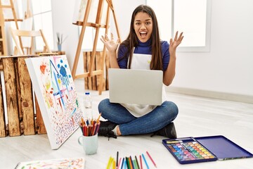 Canvas Print - Young brunette woman sitting on the floor at art studio with laptop celebrating victory with happy smile and winner expression with raised hands