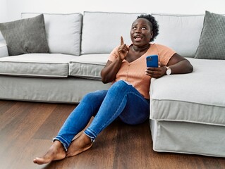 Canvas Print - Young african woman using smartphone sitting on the floor at home amazed and surprised looking up and pointing with fingers and raised arms.