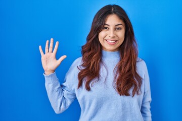 Sticker - Hispanic young woman standing over blue background showing and pointing up with fingers number five while smiling confident and happy.