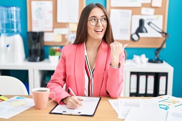 Poster - Young hispanic woman working at the office wearing glasses smiling with happy face looking and pointing to the side with thumb up.